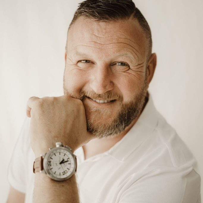 Man met een glimlach, leunend op zijn hand en een horloge om zijn pols.